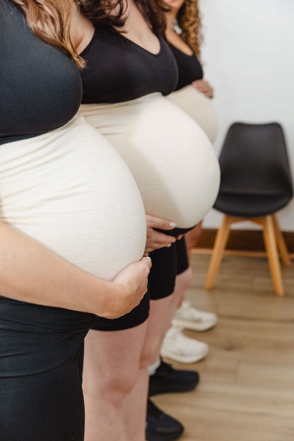 Three women wearing black tank tops and white leggings standing close together indoors, wearing abdominal support garment, tubigrip